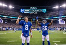 A high-energy, cinematic view of a night football game at MetLife Stadium. A rookie New York Giants quarterback, number 8, stands in the end zone with his right fist raised in triumph, smiling confidently. Beside him, a rookie Giants running back, number 26, is celebrating with both arms in the air. The bright scoreboard above them clearly shows 'GIANTS 34, EAGLES 17' in the 4th quarter. The stadium is packed with a roaring crowd waving blue and white Giants flags, celebrating a massive upset victory. Confetti is raining down, and bright stadium lights illuminate the thrilling scene